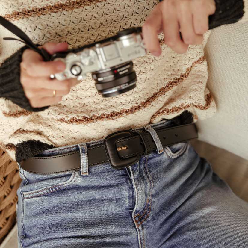 Women’s black leather belt with covered buckle, worn at the waist over jeans, camera detail - MONPIEL