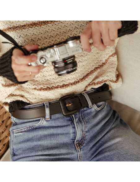 Black leather women’s belt with covered buckle worn at the waist over jeans, camera detail - MONPIEL Black leather women’s belt with covered buckle worn at the waist over jeans, camera detail - MONPIEL