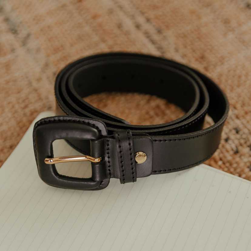 Women’s black leather belt with covered buckle, coiled on a notebook - MONPIEL