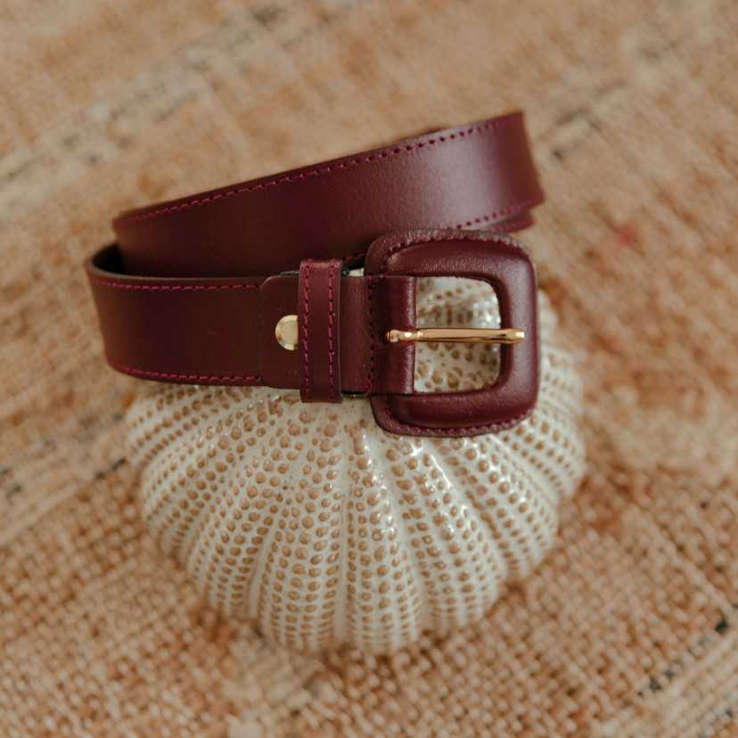 Women’s burgundy leather belt with covered buckle, coiled on a decorative object - MONPIEL