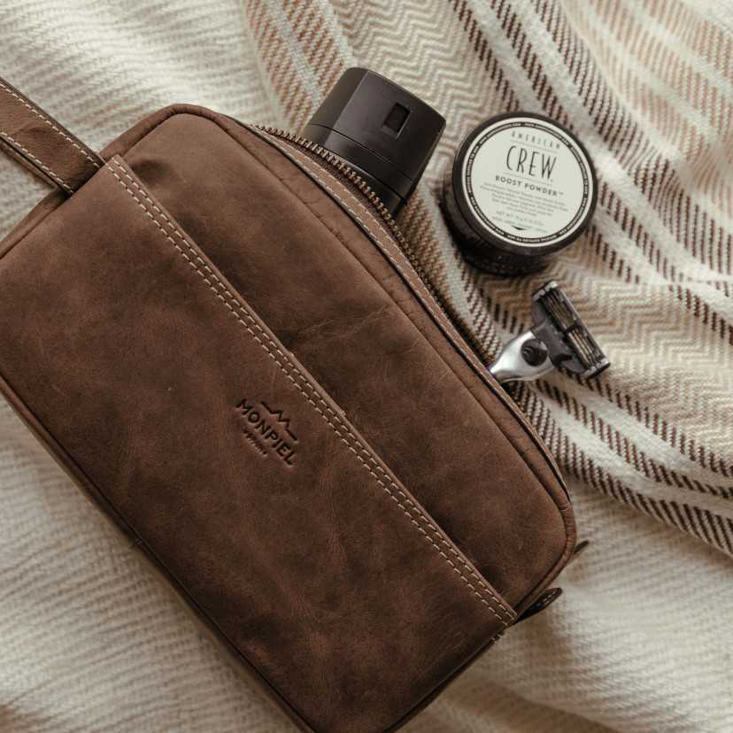 Men’s brown leather toiletry bag with front pocket, interior capacity detail, open zipper, resting on a rug - MONPIEL