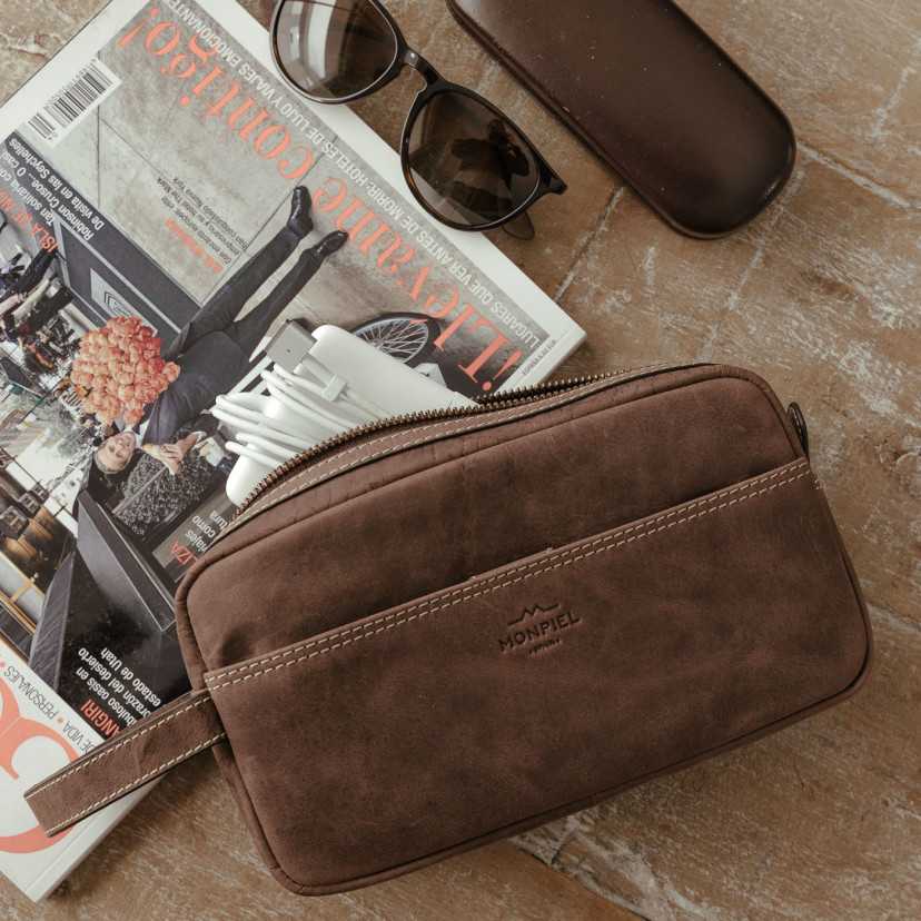 Men’s brown leather toiletry bag, front view, slightly tilted, with pocket and open zipper, resting on a magazine - MONPIEL