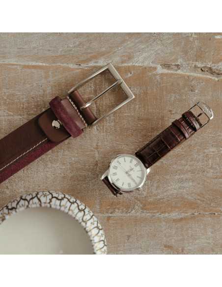 Cintura in pelle vaquetilla e serraje color bordeaux con dettaglio della fibbia accanto a un orologio su un tavolo - MONPIEL