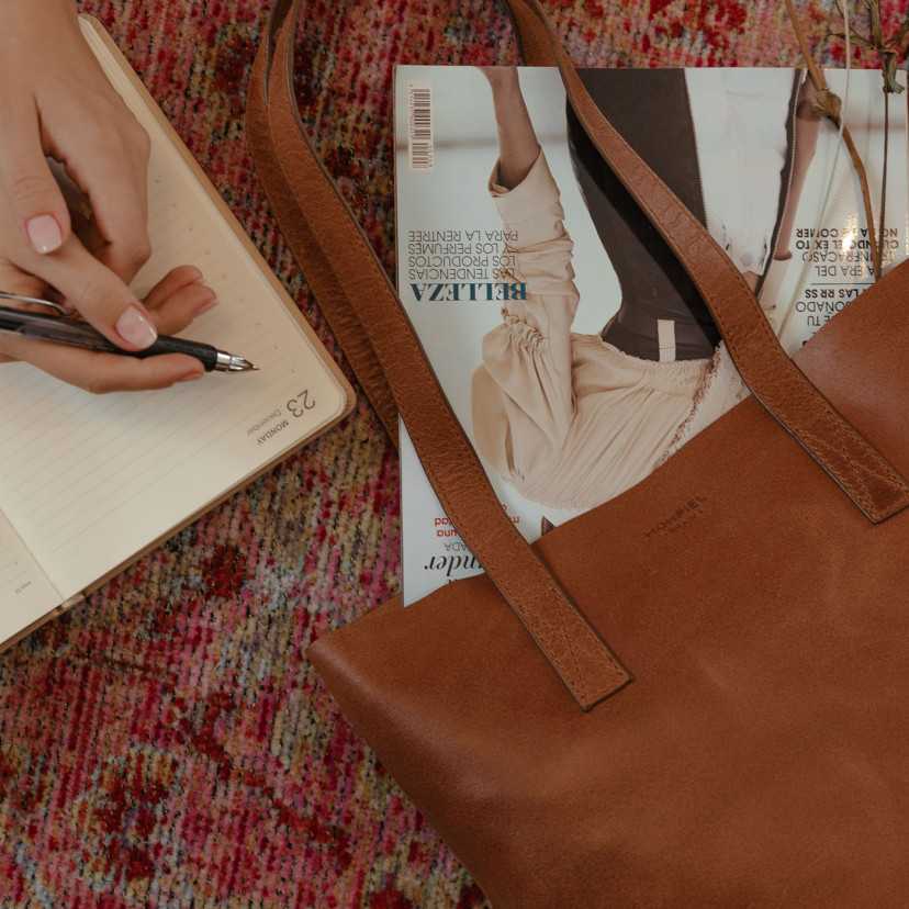 Bolsa shopper em couro com detalhe de logotipo, revista dentro, apoiada sobre um tapete - MONPIEL