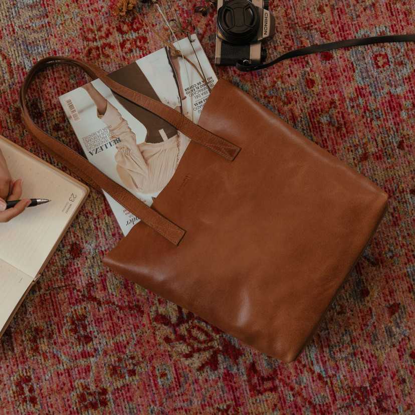 Bolsa shopper em couro, vista frontal e levemente inclinada, apoiada sobre um tapete com detalhe de câmera - MONPIEL