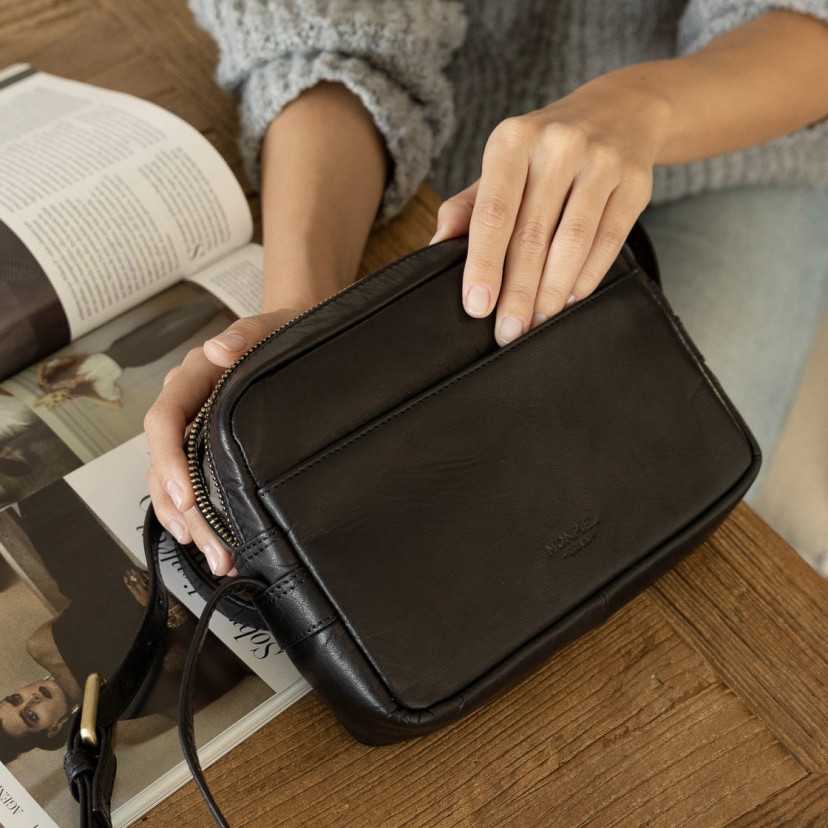 Bolsa transversal de couro preta, vista frontal levemente inclinada, apoiada sobre uma revista - MONPIEL