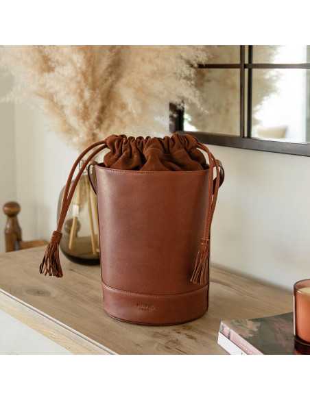 Round Havana-colored bucket bag, front view, resting on furniture with a mirror in the background - MONPIEL