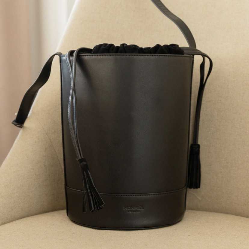 Bolsa redonda em forma de cubo de couro preto, vista frontal sobre uma poltrona - MONPIEL