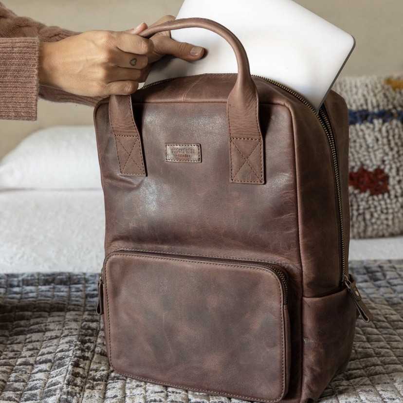 Brown leather backpack resting on the bed, placing laptop inside - MONPIEL