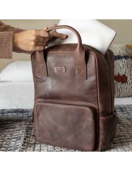 Brown leather backpack resting on a bed, laptop being placed inside - MONPIEL Brown leather backpack resting on a bed, laptop being placed inside - MONPIEL