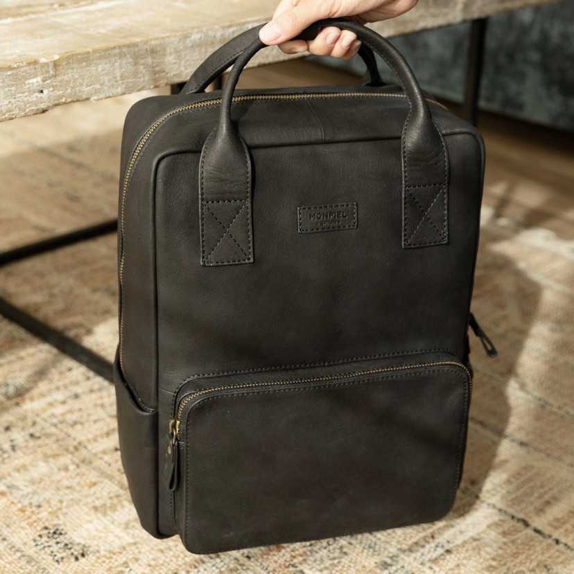 Black leather backpack held by the short handles, resting on a rug - MONPIEL