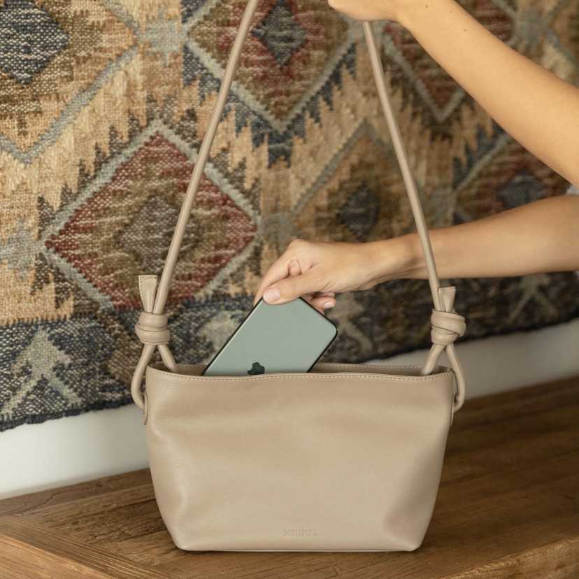 Bolsa pequena de couro em taupe com nós, vista frontal, detalhe guardando o celular - MONPIEL