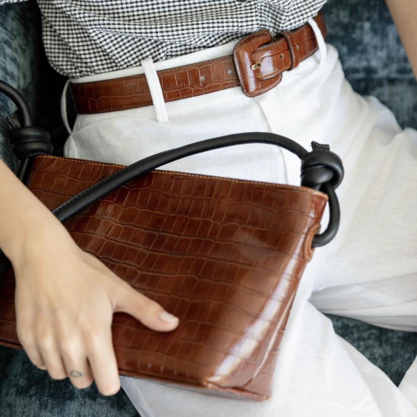 Women's belt with covered buckle and crocodile print in brown, worn over white pants, sitting while grabbing a matchin - MONPIEL