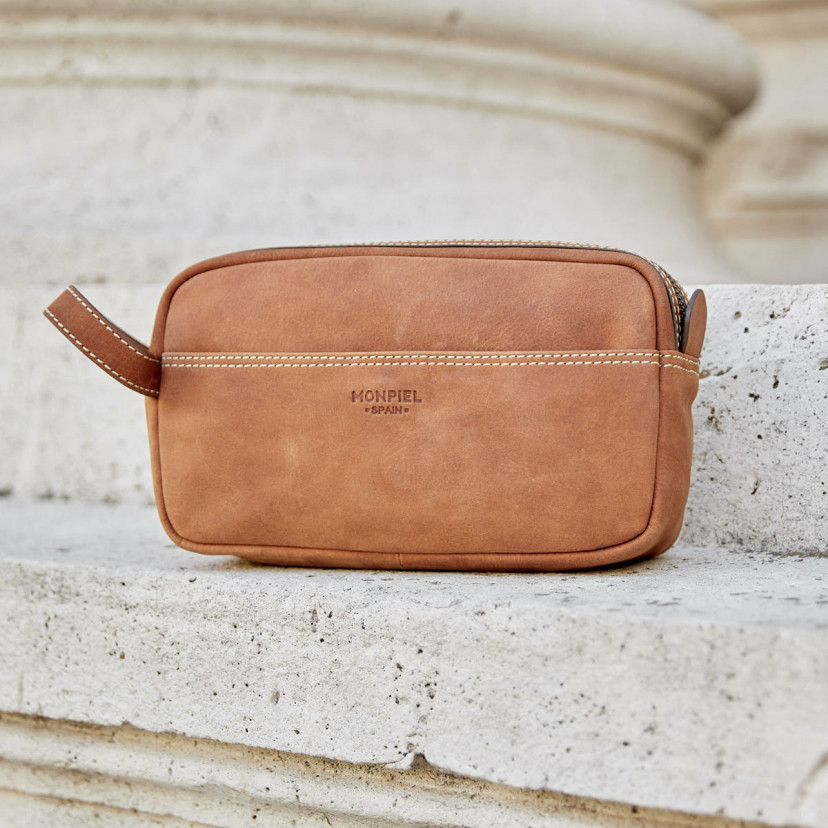 Havana leather men's toiletry bag, shown from the front with stitching detail, placed on a wall outdoors - MONPIEL