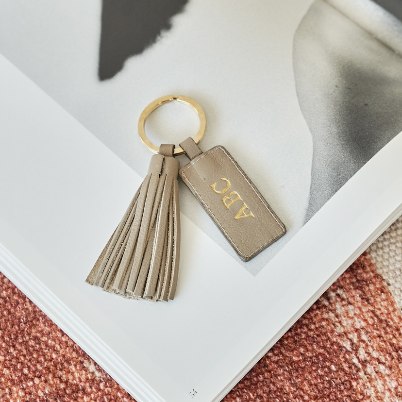 Taupe leather tassel keychain with personalized initials, placed on a magazine - MONPIEL