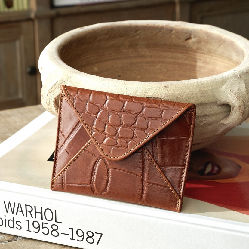 Leather envelope cardholder in tan, resting on a book and ceramic piece - MONPIEL