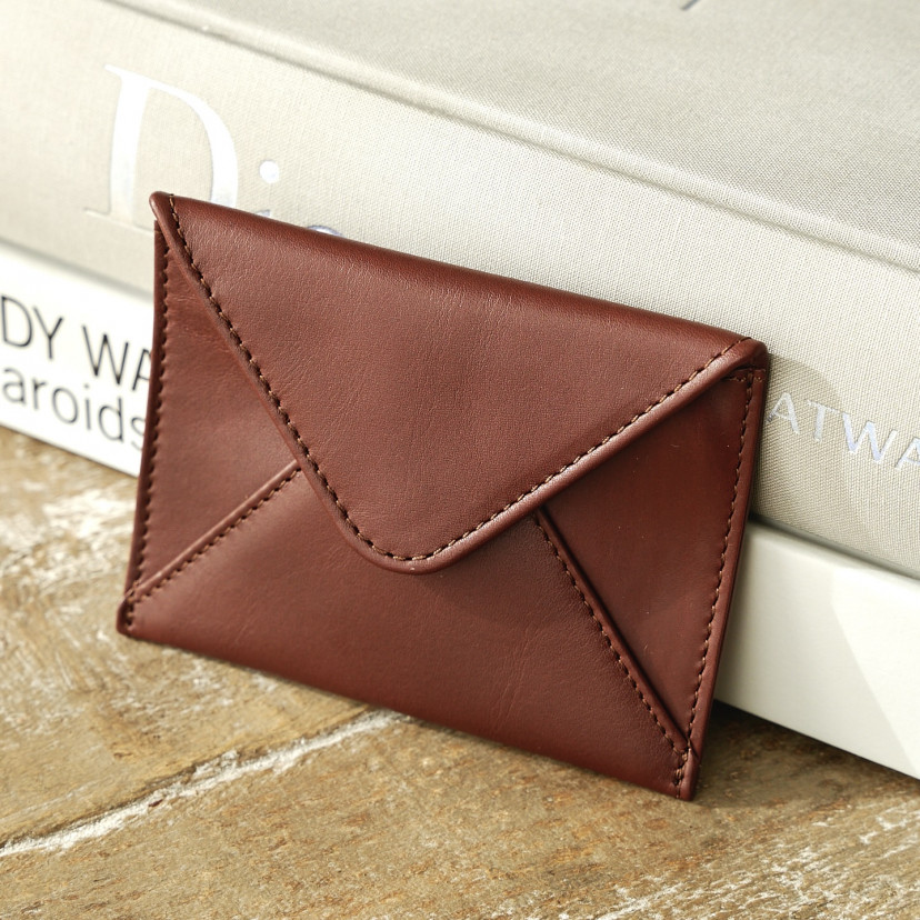 Leather envelope cardholder in brown, resting on books on a wooden table - MONPIEL