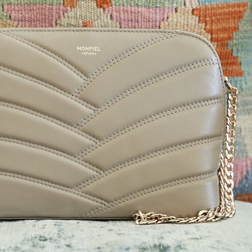 Bolsa acolchoada de couro com corrente dourada na cor taupe em primeiro plano com detalhe do logo apoiado sobre almofa - MONPIEL