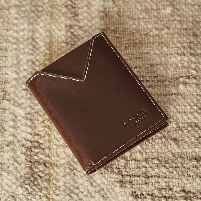 Small brown leather men's wallet, shown from the front at an angle, resting on a carpet - MONPIEL