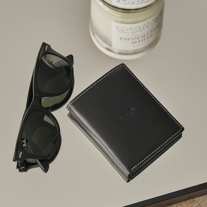Simple black leather billfold with central logo, resting on a mirror with sunglasses beside it - MONPIEL