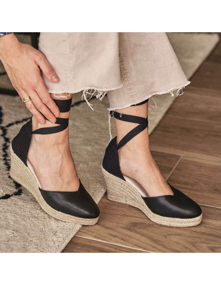 Elba Keilabsatz-Espadrille aus schwarzem Leder, Füße auf einem Teppich, Seitenansicht - MONPIEL Elba Keilabsatz-Espadrille aus schwarzem Leder, Füße auf einem Teppich, Seitenansicht - MONPIEL
