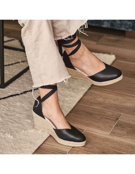 Elba Keilabsatz-Espadrille aus schwarzem Leder, Beine überkreuzt, Seitenansicht - MONPIEL Elba Keilabsatz-Espadrille aus schwarzem Leder, Beine überkreuzt, Seitenansicht - MONPIEL