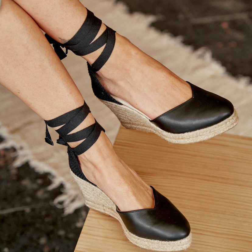 Elba wedge espadrille in black leather, legs crossed, foot resting on the table - MONPIEL