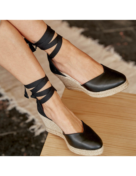 Espadrille à talon compensé Elba en cuir noir, jambes croisées avec un pied posé sur la table - MONPIEL