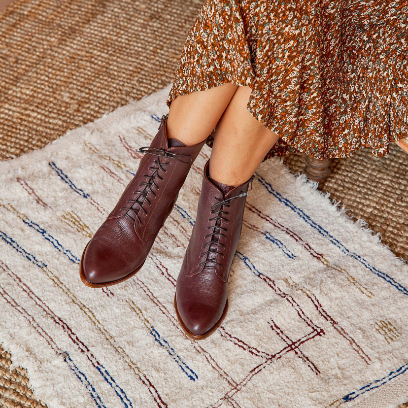 Women’s leather pointed-toe ankle boots in burgundy color, legs crossed, resting on a rug - MONPIEL