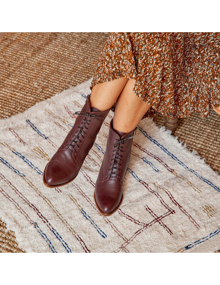 Women’s leather pointed-toe ankle boots in burgundy color, legs crossed, resting on a rug - MONPIEL Women’s leather pointed-toe ankle boots in burgundy color, legs crossed, resting on a rug - MONPIEL