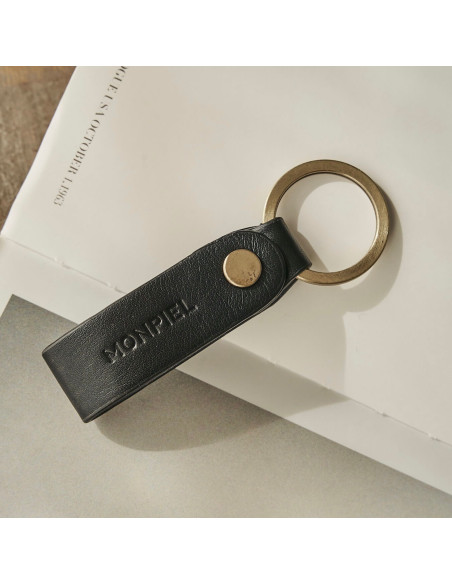 Unisex leather keychain in black color, slightly tilted, resting on a magazine - MONPIEL Unisex leather keychain in black color, slightly tilted, resting on a magazine - MONPIEL