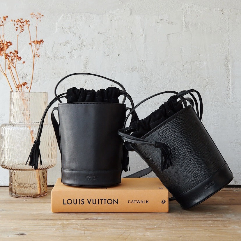 Small round bucket leather bag in black color resting on a book, alongside a small black snakeskin bucket bag - MONPIEL