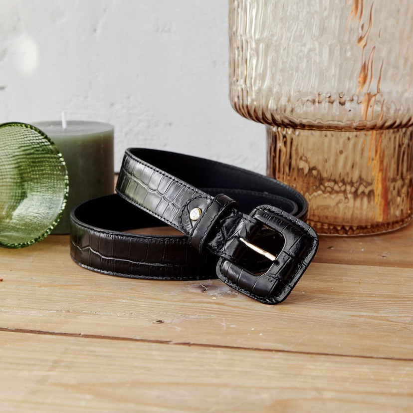 Women’s covered buckle belt with crocodile pattern in black, coiled next to a candle and a vase - MONPIEL