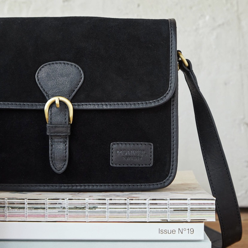 Bolsa satchel com fivela em camurça preta, vista frontal apoiada em livros, detalhe do logotipo - MONPIEL