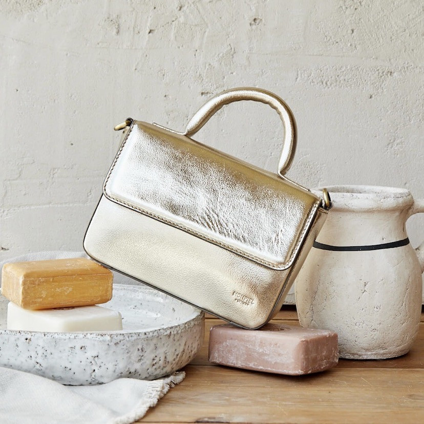 Bolsa pequena estruturada de couro metálico champanhe, inclinada apoiada sobre sabão, ao lado de um vaso - MONPIEL