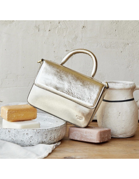 Bolsa pequena estruturada de couro metálico champanhe, inclinada apoiada sobre sabão, ao lado de um vaso - MONPIEL Bolsa pequena estruturada de couro metálico champanhe, inclinada apoiada sobre sabão, ao lado de um vaso - MONPIEL