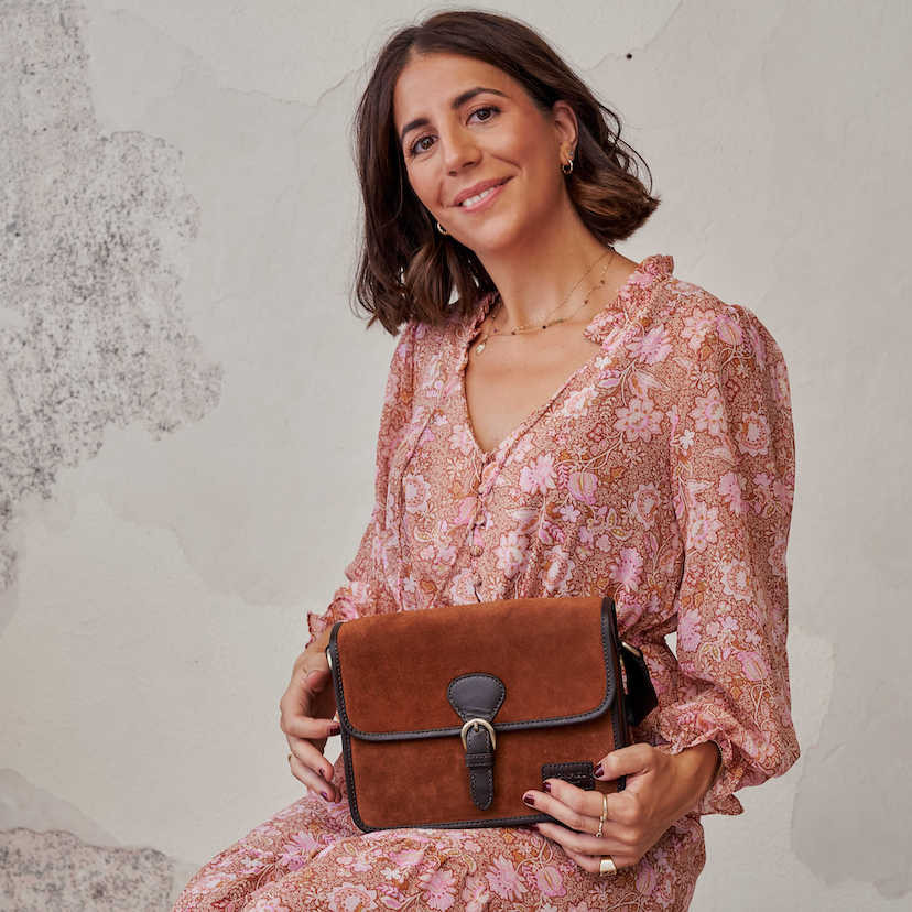 Bolsa satchel em camurça marrom com fivela, combinada com um vestido, segurada com as mãos - MONPIEL