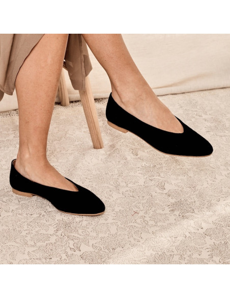 Black suede flat ballerina, legs crossed, sitting in profile with a carpet in the background - MONPIEL Black suede flat ballerina, legs crossed, sitting in profile with a carpet in the background - MONPIEL