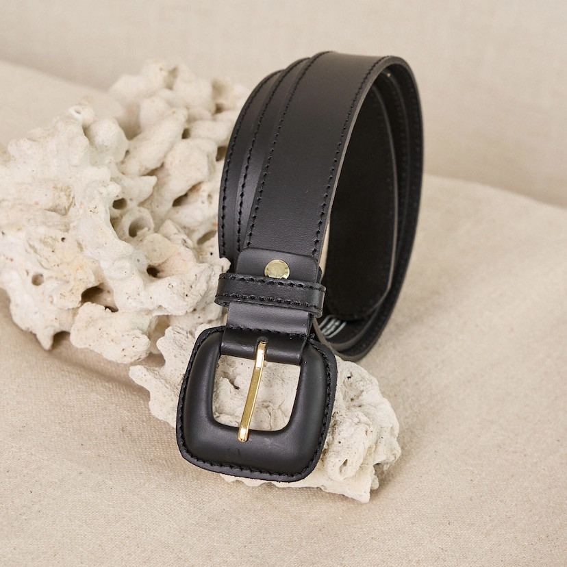 Black leather women's belt with covered buckle, coiled, with a sofa in the background - MONPIEL