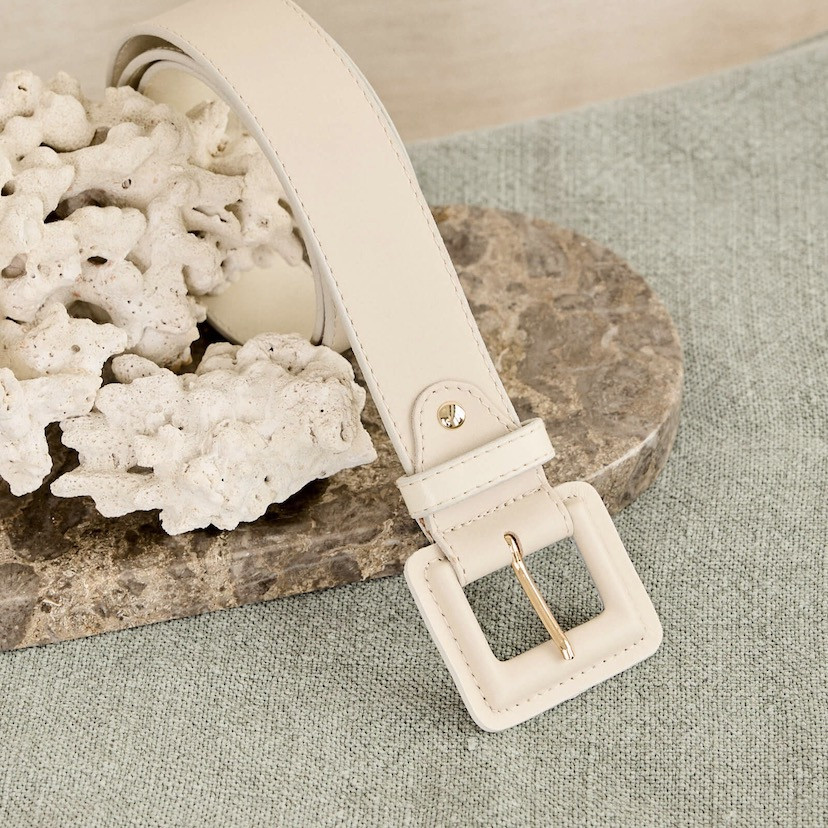 Wide women's leather belt with covered buckle in beige, coiled on marble with focus on the buckle - MONPIEL