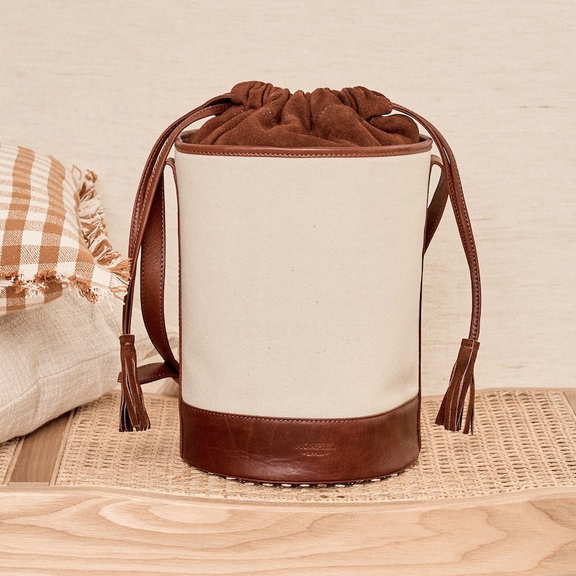 Bolsa cubo de lona e couro em cor couro, vista frontal, apoiada sobre banco com almofadas ao lado - MONPIEL