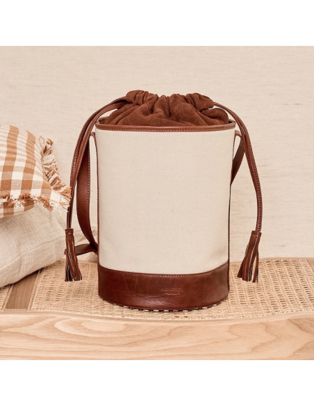 Borsa cubo in canvas e pelle color cuoio, vista frontale, appoggiata su una panca con cuscini accanto - MONPIEL