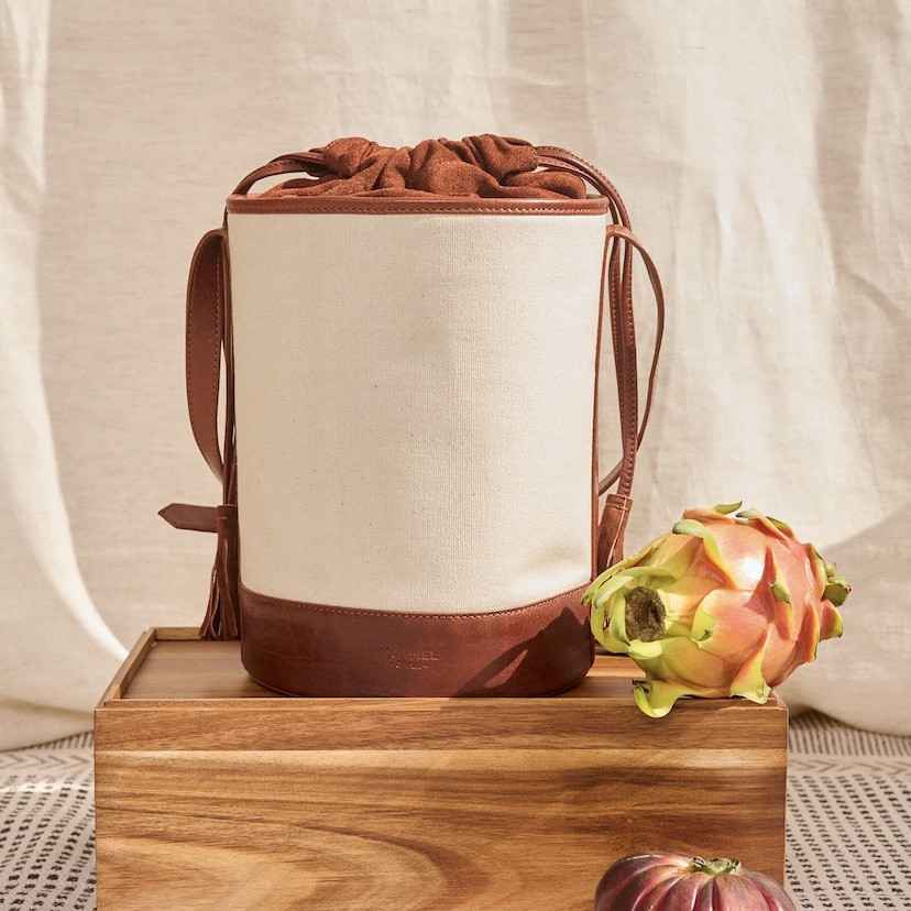 Bolsa cubo em canvas e couro em tom couro, vista frontal apoiada em madeira com cortina ao fundo - MONPIEL