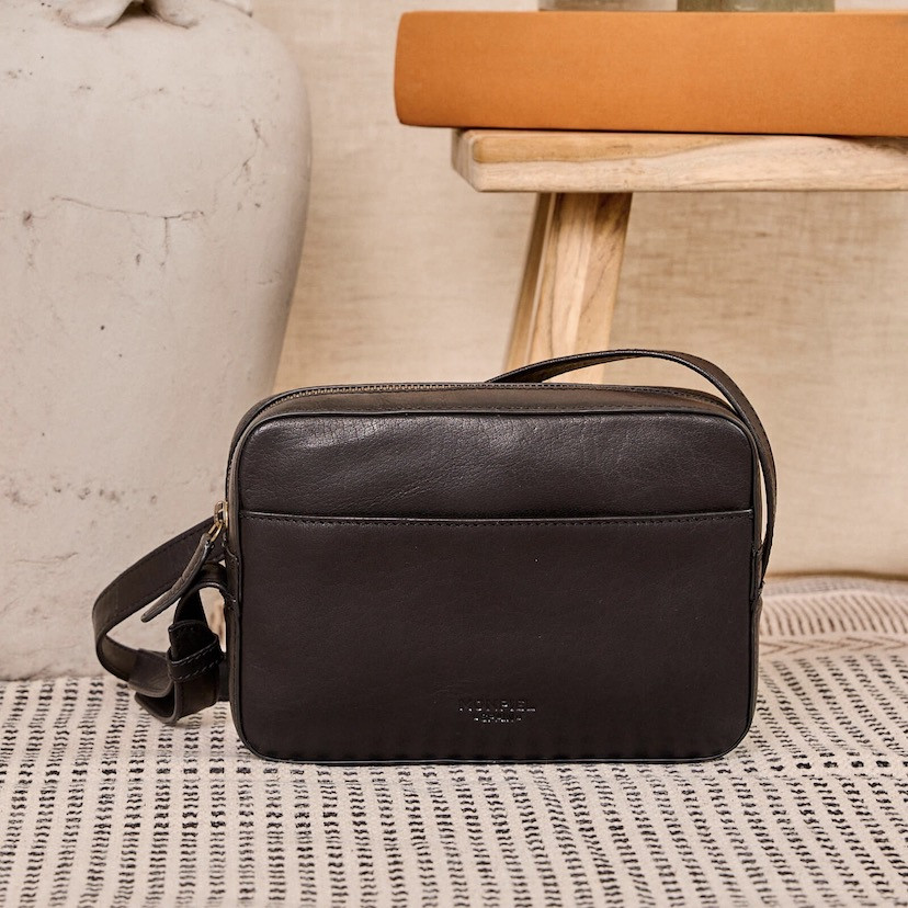 Bolsa tiracolo de couro em preto, vista frontal apoiada sobre um tapete com uma cadeira ao fundo - MONPIEL