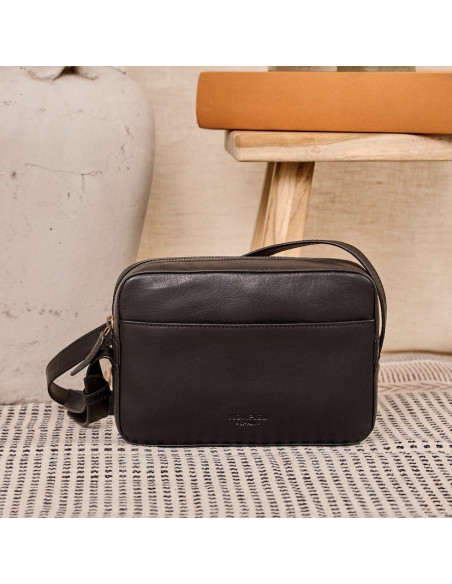 Black leather crossbody bag, front view, resting on a carpet with a chair in the background - MONPIEL Black leather crossbody bag, front view, resting on a carpet with a chair in the background - MONPIEL