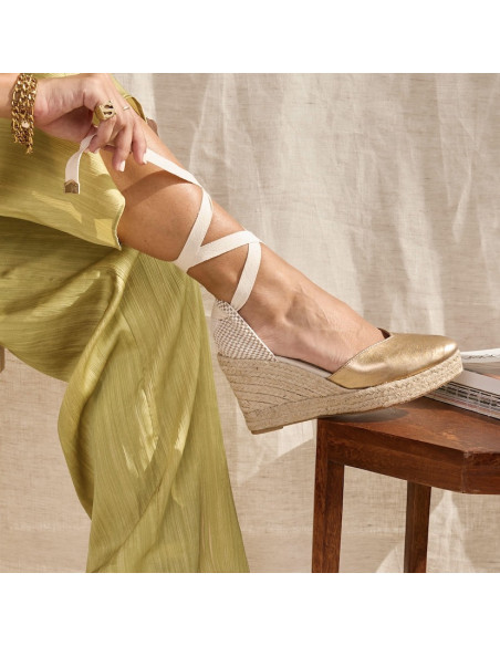 Espadrille com cunha Elba de couro dourado, pé apoiado em uma mesinha, detalhe da amarração - MONPIEL Espadrille com cunha Elba de couro dourado, pé apoiado em uma mesinha, detalhe da amarração - MONPIEL