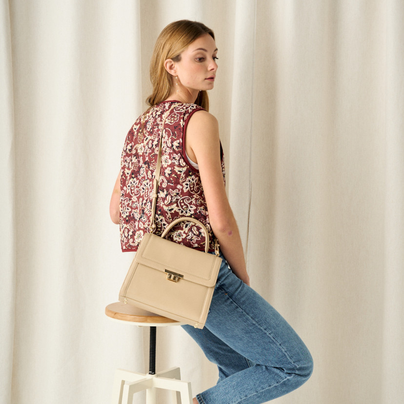 60's leather handbag for women in beige, front view hanging on shoulder, medium shot of model seated on stool - MONPIEL