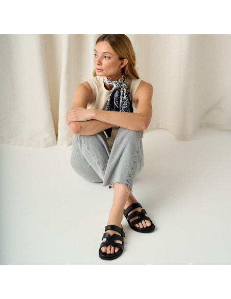 Lucía Bio-Sandale in Schwarz, Gesamtaufnahme des Modells, das auf dem Boden sitzt und die Beine übereinanderschlägt - MONPIEL