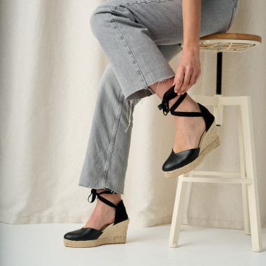 Espadrille à talon compensé Elba en cuir noir, jambes croisées avec un pied posé sur la table - MONPIEL 2