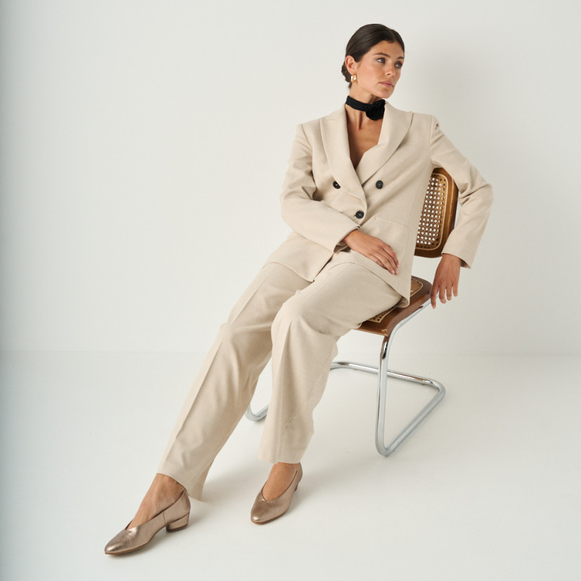 Heeled flat slipper in cappuccino bronze with full shot of the model sitting on the chair, perspective view - MONPIEL